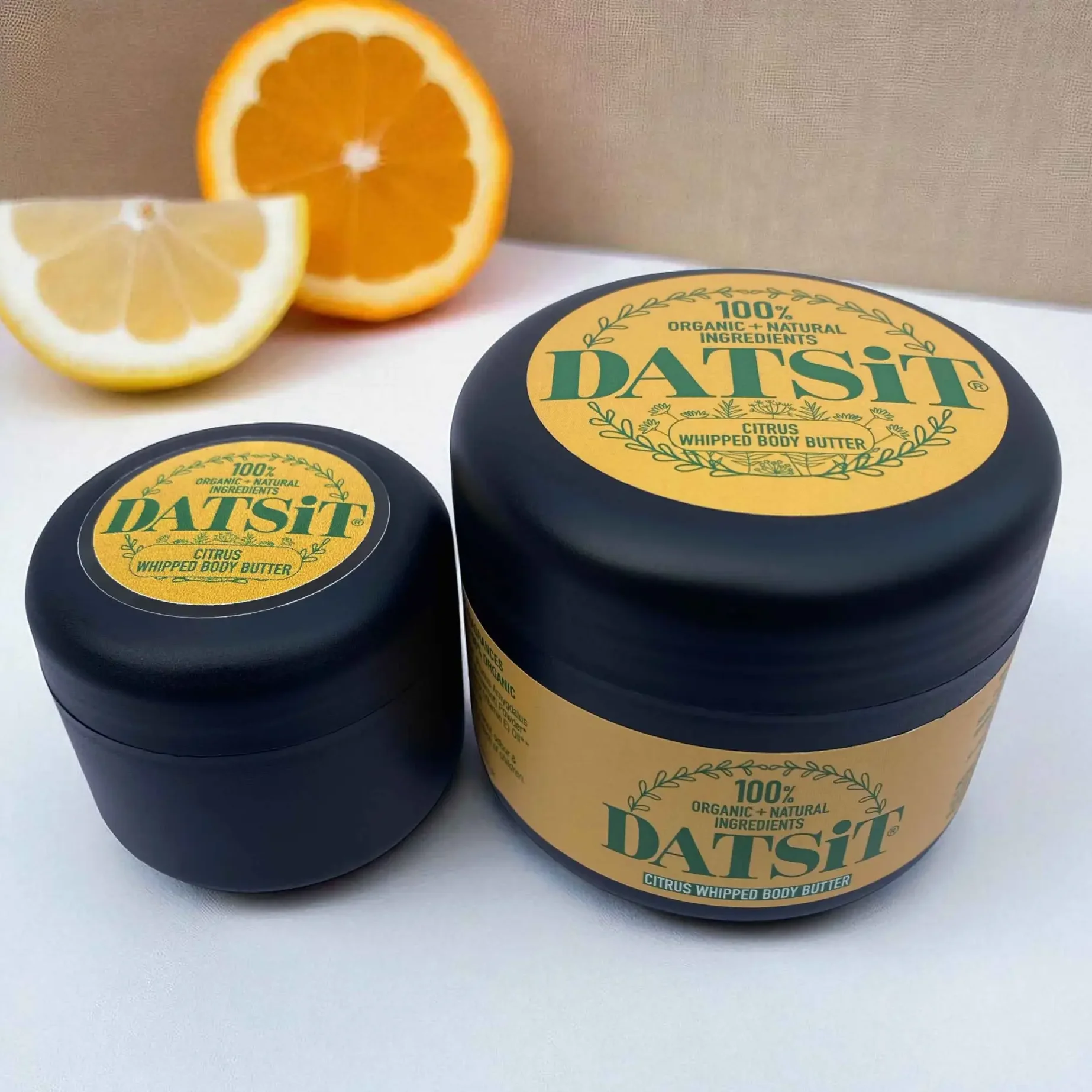 Two jars of Datsit organic citrus whipped body butter, one 50ml jar, and one 150ml jar on a muted background with soft shadows and a slice of orange and lemon in the background