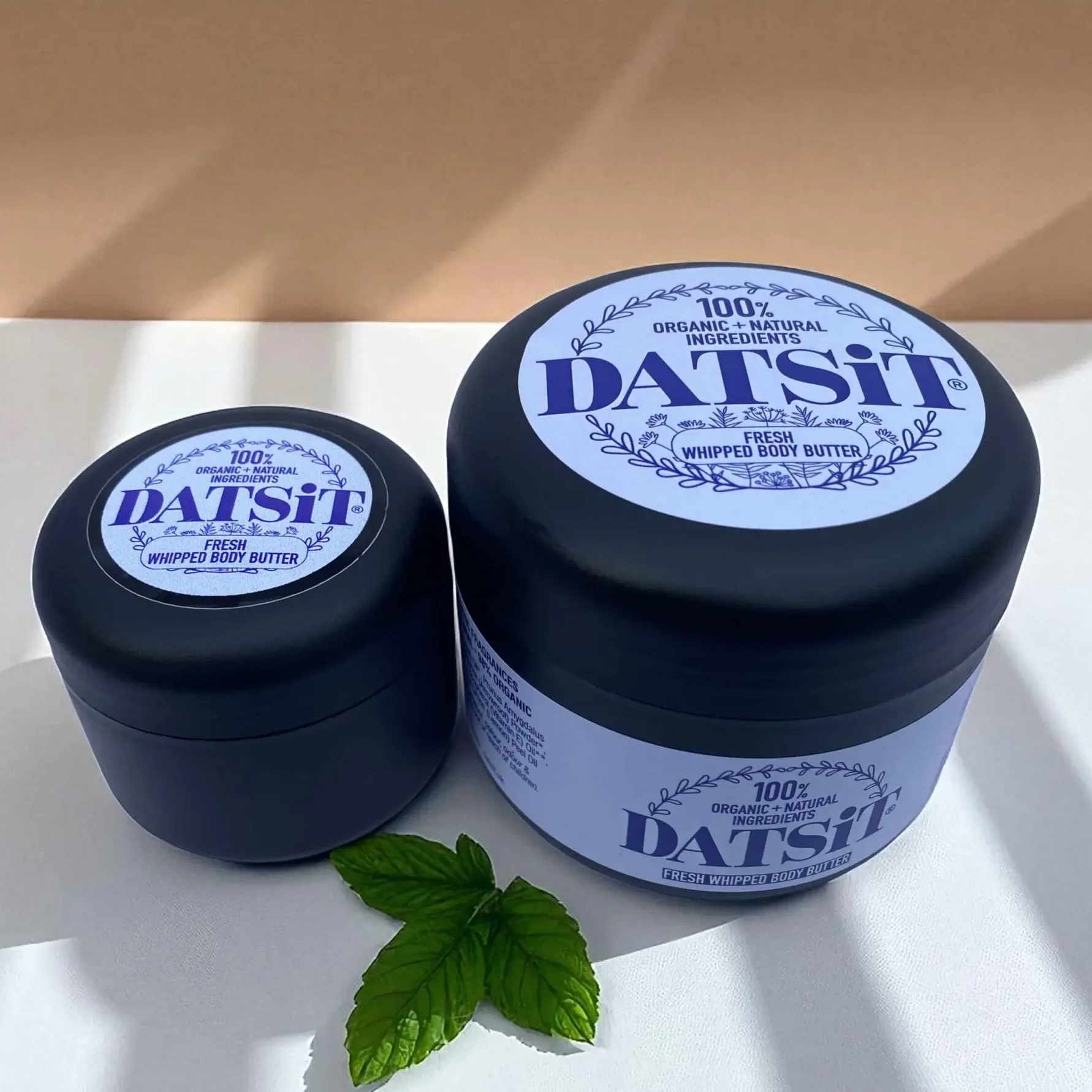 Two jars of Datsit organic fresh whipped body butter, one 50ml jar, and one 150ml jar on a muted background with soft shadows, with a few mint leaves in the foreground