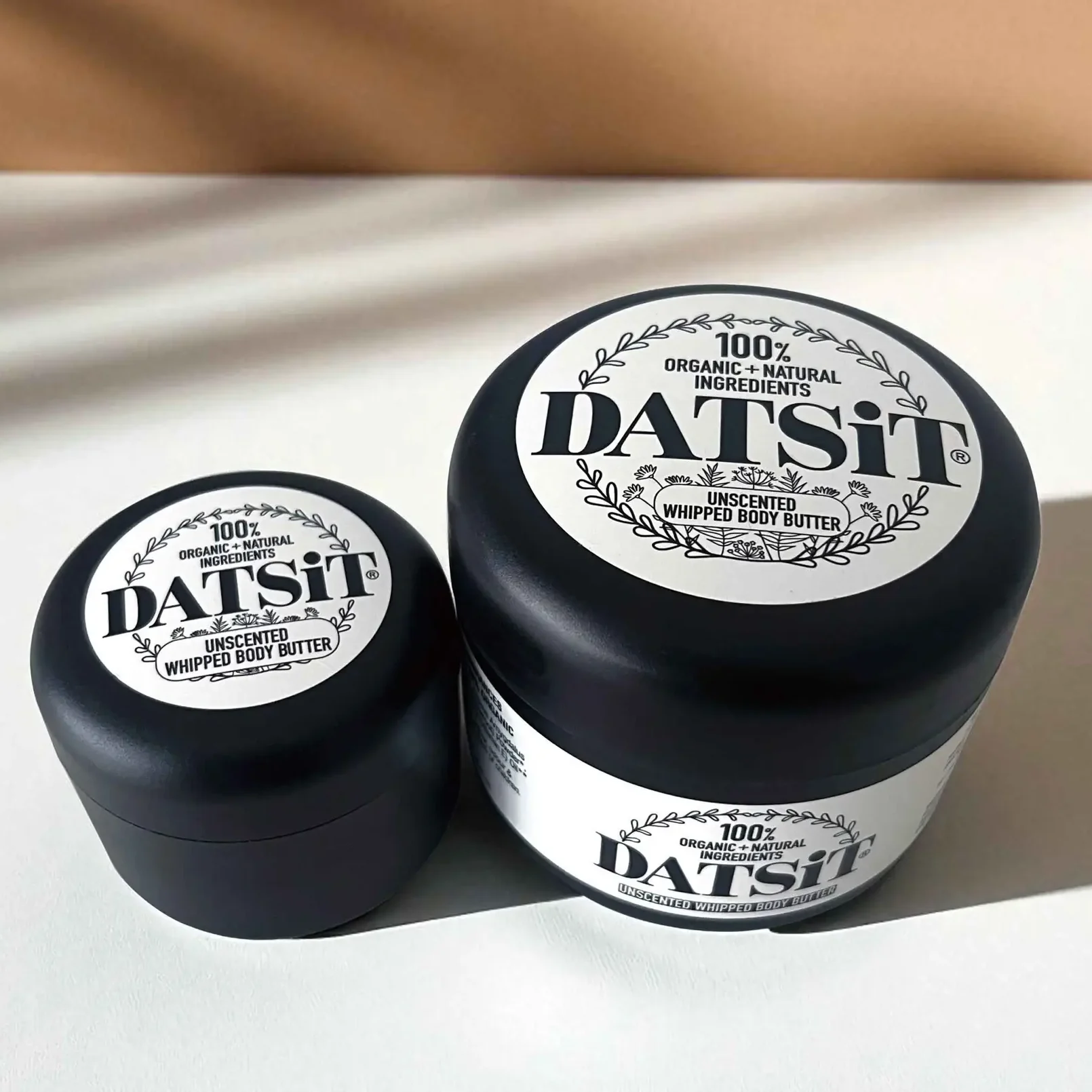 Two jars of Datsit organic unsceneted whipped body butter, one 50ml jar, and one 150ml jar on a muted background with soft shadows